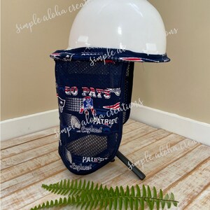 Pu&ograve; includere: Vista laterale di un parasole per il collo blu e bianco con tessuto a rete e un casco rigido bianco in cima. Il parasole per il collo ha un tema dei New England Patriots con le parole "Go Pats" e "Patriots" stampate su di esso. Il parasole per il collo &egrave; seduto su una superficie di legno con una pianta di felce verde in primo piano.