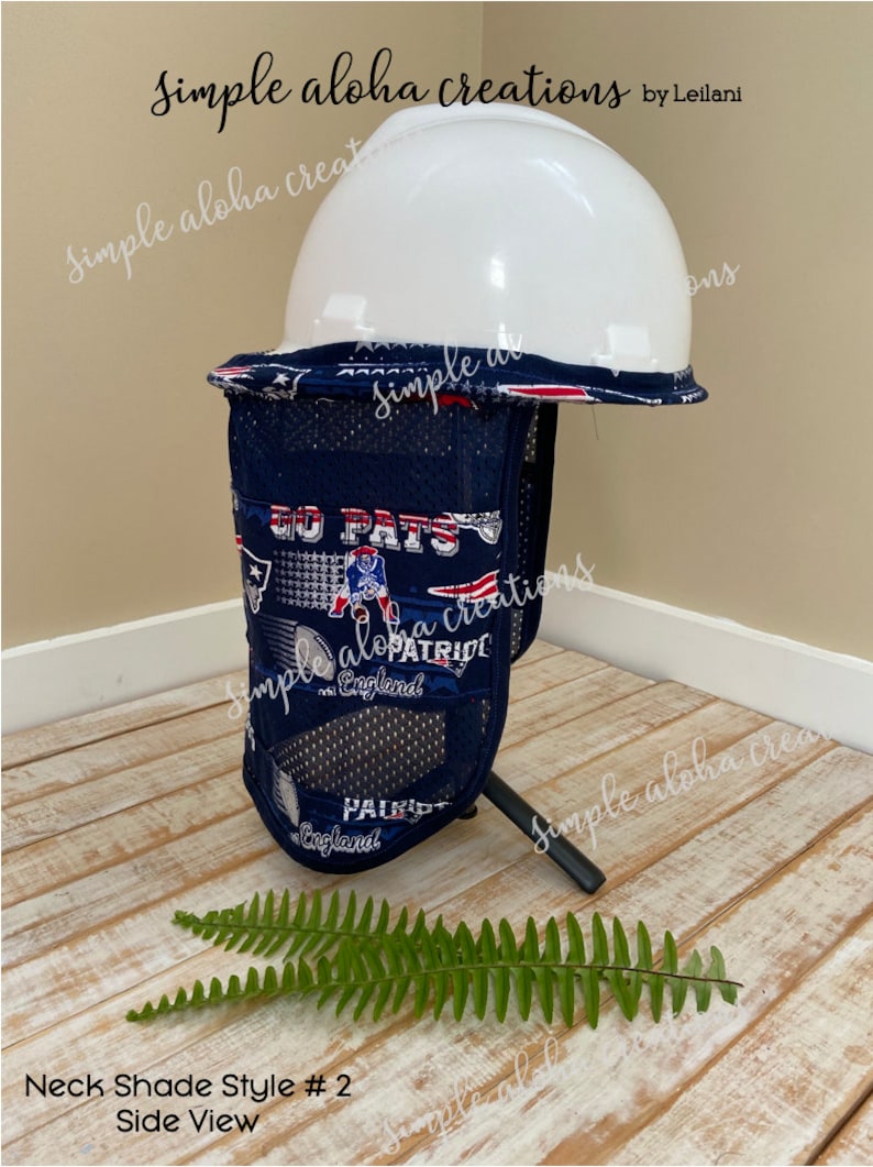 Pu&ograve; includere: Vista laterale di un parasole per il collo blu e bianco con tessuto a rete e un casco rigido bianco in cima. Il parasole per il collo ha un tema dei New England Patriots con le parole "Go Pats" e "Patriots" stampate su di esso. Il parasole per il collo &egrave; seduto su una superficie di legno con una pianta di felce verde in primo piano.