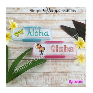 May include: Two clear plastic lunch boxes with colorful designs and the word "Aloha" on them. The lunch boxes are on a wooden surface with flowers and leaves around them.