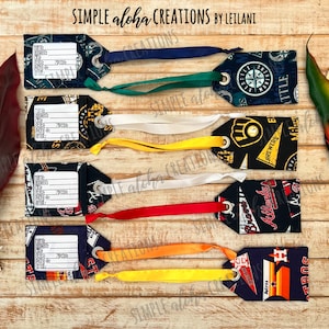 May include: Six fabric luggage tags with different baseball team logos. The tags are navy blue with white, red, yellow, and orange accents. Each tag has a white label with a blank space for writing information. The tags are attached to white, yellow, red, and green ribbons.