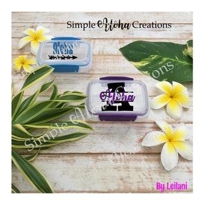 May include: Two clear plastic containers with lids. One container has a blue lid and the other has a purple lid. Both containers have a design on them that says "Aloha" in black and purple lettering. The containers are on a wooden surface with white flowers.