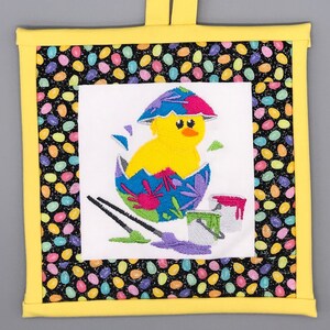 Puede incluir: Un pollito amarillo asomándose de un huevo de Pascua colorido con pinceles y botes de pintura. El pollito está rodeado de un fondo negro con patrones coloridos en forma de judías. La imagen está enmarcada con un borde amarillo.