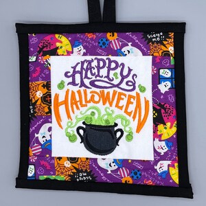 May include: A purple and black fabric wall hanging with a white square in the center. The square has an embroidered design that says "Happy Halloween" with a cauldron and green potion. The fabric is decorated with ghosts, bats, and other Halloween imagery.