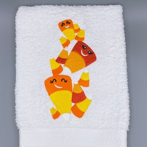 Può includere: Asciugamano bianco ricamato con personaggi di caramelle di mais sorridenti in giallo, arancione e rosso. L'asciugamano è realizzato in un materiale morbido e assorbente, perfetto per la stagione di Halloween. Il design delle caramelle di mais aggiunge un tocco festoso.