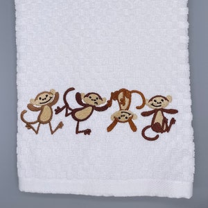 May include: White kitchen towel with brown embroidered monkeys. The monkeys are in a line and are all facing different directions.