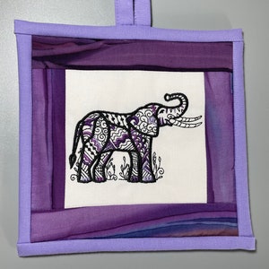 Może przedstawiać: Wiszący obraz z fioletowej tkaniny z białym kwadratem w środku, na którym znajduje się haftowany czarny słoń z misternymi wzorami. Obraz ma fioletową pętlę z tkaniny u góry do zawieszenia.