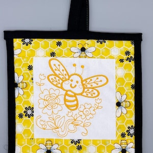 以下が含まれることがあります： 白く刺繍された蜂と花がついた、黄色と黒の蜂の鍋つかみ。鍋つかみには黒い吊り下げループが付いています。