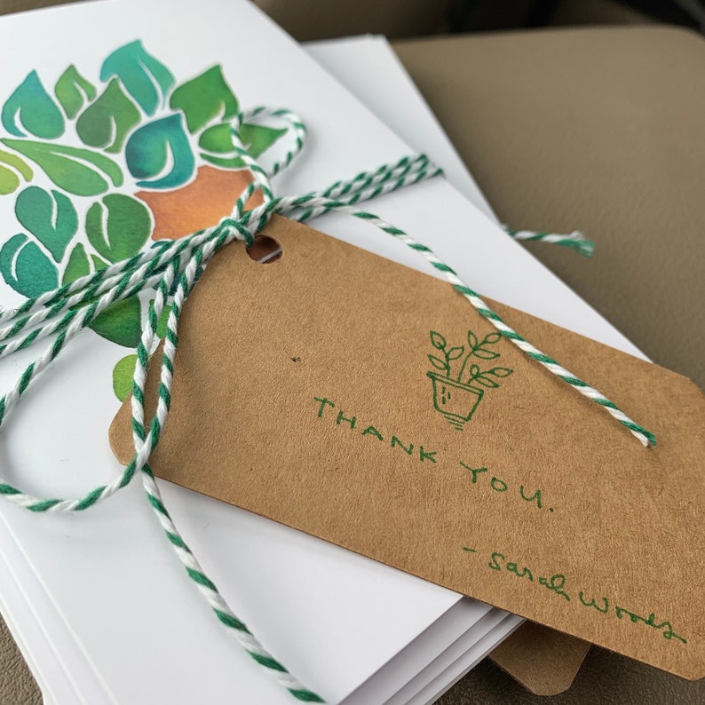 May include: A stack of blank greeting cards with a green and orange watercolour design. The cards are tied together with twine and a brown tag that says "THANK YOU."  The tag also has a small green drawing of a potted plant.