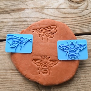 May include: Three blue rubber stamps with insect designs. The stamps are on a brown clay surface. The designs are a bee, a fly, and a wasp.