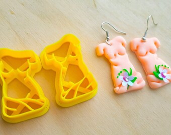 May include: A pair of handmade, flesh-toned polymer clay earrings in the shape of a torso, adorned with floral accents. Two yellow plastic cutters in the same shape are also present. The earrings have silver-colored ear wires.