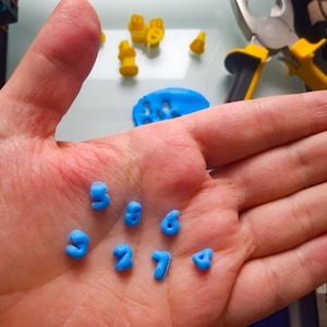 May include: A hand holding a collection of blue plastic number molds. The numbers include 3, 8, 6, 3, 2, 7, and 4.