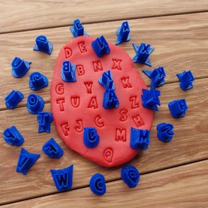 May include: A red play dough mat with the alphabet embossed on it. Blue plastic alphabet cookie cutters are scattered around the mat.