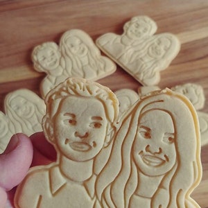 May include: A light-colored, flat cookie shaped like a couple, featuring embossed facial features and hair details. The cookie depicts a person with short hair next to a person with long, wavy hair. Several identical cookies are arranged in the background.