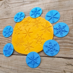 May include: A selection of blue snowflake stamps and a yellow clay surface with snowflake imprints. The stamps are positioned around the clay, displaying different snowflake patterns. The wooden surface provides a rustic backdrop.