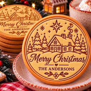 May include: A round, light brown cookie featuring an embossed winter scene with a house, pine trees, and a star. The text reads Merry Christmas and THE ANDERSONS, with a small heart and floral accents centered below the text.
