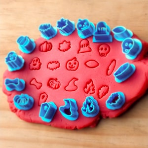 May include: A set of blue plastic Halloween-themed cookie cutters on a red play dough surface. The cutters include shapes like ghosts, pumpkins, bats, witches' hats, and more.
