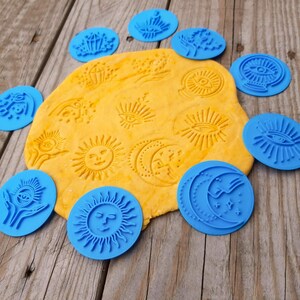 以下が含まれることがあります： 太陽、月、目、クリスタルなどの様々な天体や神秘的なデザインが施された円形の青いプラスチック製スタンプのセット。中央には、これらの模様が型押しされた黄色い生地が置かれています。