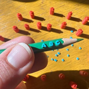 May include: A green pencil with the letters "ABC" in blue is held. Small red and blue objects, possibly stamps or erasers, are scattered on a yellow surface. The image is brightly lit, suggesting a craft or educational theme.