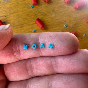 Könnte beinhalten: Nahaufnahme einer Hand, die den Namen "NOAH" in kleinen, türkisfarbenen Buchstaben hält. Kleine rote zylindrische Objekte und türkisfarbene Punkte sind auf einer gelben Holzoberfläche verstreut.