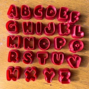 May include: A complete set of twenty-six red plastic alphabet cookie cutters arranged in rows. Each individual cutter represents a letter from A to Z, featuring a thick, molded frame designed for cutting dough into specific letter shapes.