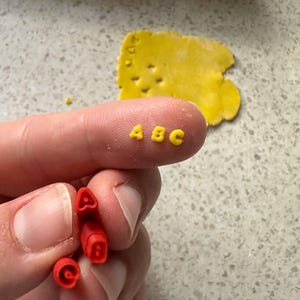 May include: A close-up of yellow ABC letters and red letter stamps. The letters are on a fingertip, with a yellow dough shape in the background. The red stamps are held in the hand. The letters are for baking or crafting.