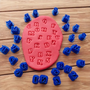 May include: A set of 26 blue plastic alphabet cookie cutters on a wooden surface. The cutters are arranged around a red playdough with the letters of the alphabet pressed into it.