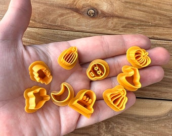 May include: A collection of orange plastic Halloween-themed cookie cutters, including pumpkins, a skull, a bell, a crescent moon, and a star. The cutters are held in a hand against a wooden background.