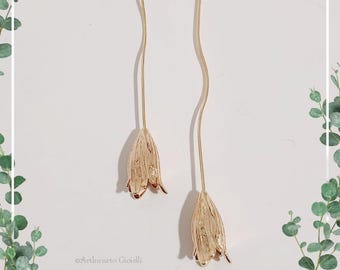 May include: A pair of gold-colored drop earrings. Each earring features a hexagonal stud, a thin, wavy stem, and a textured, tulip-shaped pendant. The earrings are displayed on a white background with green foliage accents.