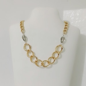 Könnte beinhalten: Goldfarbene Kette mit großen, ineinandergreifenden kreisförmigen Gliedern. Die Halskette hat auf jeder Seite eine kleinere Kette, die mit einer grauen, tropfenförmigen Perle verbunden ist. Die Halskette wird auf einer weißen Schaufensterpuppe präsentiert.