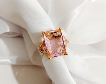 May include: A gold ring with a large, square-shaped pink gemstone. The ring has smaller, clear gemstones on either side of the main stone.