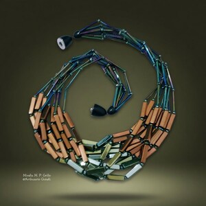 Può includere: Una collana a più fili con perline rettangolari allungate in tonalità metalliche rame, bronzo e blu-verde iridescente. I fili sono raccolti a ciascuna estremità da chiusure coniche nere. Il testo Mirella M. P. Grillo Artincarto Gioielli è visibile.