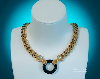 Könnte beinhalten: Eine goldfarbene Kettenhalskette mit dicken, ineinandergreifenden Panzerkettengliedern. Die Kette endet in zwei stilisierten Metallköpfen, die einen glatten, kreisförmigen schwarzen Ringanhänger in der Mitte halten. Der Text "Movilla M. P. Grillo ©Artigianato Gioielli" ist sichtbar.