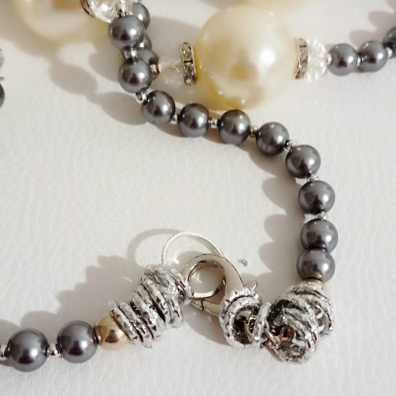 May include: A grey pearl necklace with a silver clasp and two large white pearls. The necklace is made of small, round pearls and has a simple design.