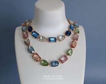 Può includere: Una collana a doppio filo con cristalli sfaccettati rettangolari e a goccia nelle tonalità del blu, verde e rosa, montati su una catena in metallo dorato. Il testo Mirella M.P. Grillo ArtIncarto Gioielli è stampato sotto la collana.