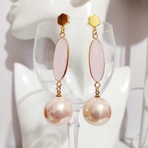 May include: A pair of dangle earrings with a gold-tone finish. Each earring features a hexagonal stud, a light pink oval, and a large, pearlescent pink sphere. The earrings are suspended from a delicate chain and are displayed against a clear glass.