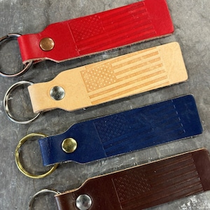 May include: Four leather keychains with a US flag design. The keychains are red, tan, blue, and brown. Each keychain has a silver key ring.