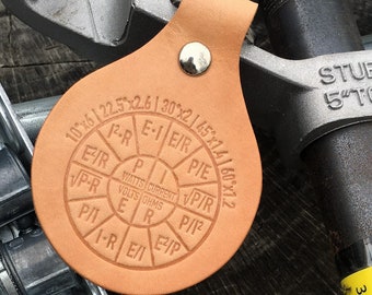 May include: A tan leather tag featuring a circular electrical power formula wheel with P, I, E, and R variables, along with various mathematical equations and degree measurements. The tag is secured with a silver metal rivet and hangs from a metal tool.
