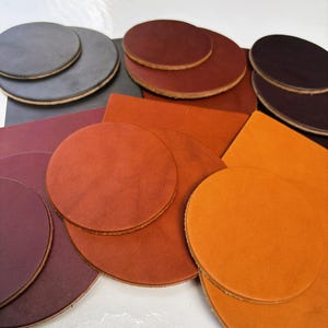 Puede incluir: Una colección de piezas de cuero circulares y cuadradas en varios colores, incluyendo negro, marrón oscuro, burdeos, naranja óxido y naranja brillante. Los artículos de cuero tienen superficies lisas y bordes visibles, dispuestos en capas.