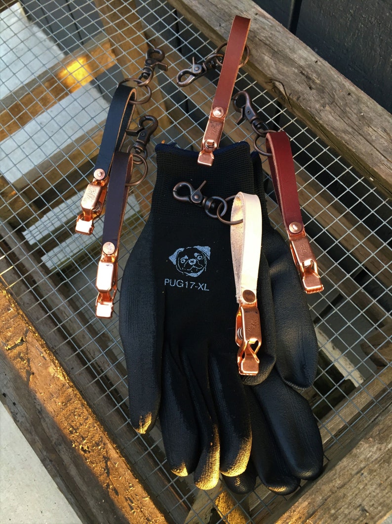Puede incluir: Cinco llaveros de cuero con clips y anillos de cobre. Los llaveros est&aacute;n sobre un guante de trabajo negro con el texto "PUG 17-XL" impreso en el dorso de la mano.