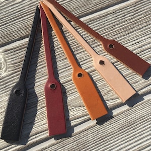 Puede incluir: Cinco etiquetas de equipaje de cuero en varios colores: negro, burdeos, naranja, beige y marrón. Cada etiqueta tiene una base rectangular con una parte superior redondeada y un pequeño agujero para la fijación. Las etiquetas están dispuestas sobre una superficie de madera clara.
