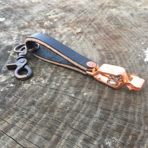 Puede incluir: Un llavero de cuero negro con un clip de cobre y un anillo de llave de cobre.
