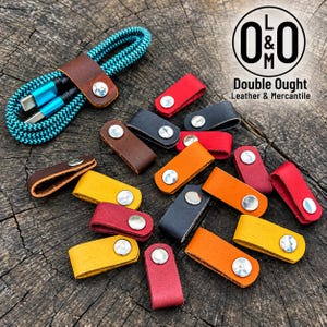 Puede incluir: Una colección de pequeños organizadores de cables de cuero en rojo, naranja, amarillo, marrón y negro, cada uno con un botón de presión de metal plateado. Un organizador envuelve un cable de carga trenzado azul y negro. El logo dice O&M Double Ought Leather & Mercantile.