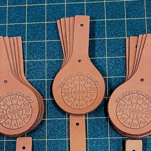 May include: Several brown leather teardrop-shaped pieces with a hole at the top. Each piece has a circular design with text and measurements like 10"x6 and 22.5"x2.6. The pieces are arranged on a green grid.