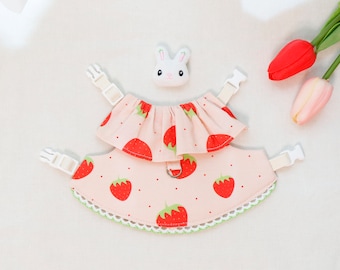 Puede incluir: Un arnés para perro rosa y blanco con estampado de fresas y ribete blanco. El arnés tiene dos correas ajustables con hebillas de plástico blanco y un anillo de metal blanco.