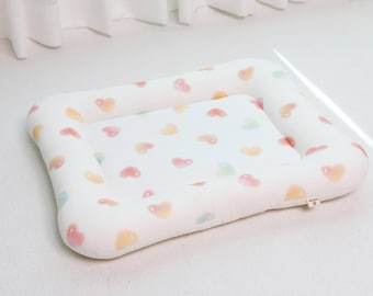 May include: A rectangular pet bed with a white base and a soft, cushioned design. The bed is adorned with a pattern of pastel-colored hearts in shades of pink, yellow, and green. The bed has a raised, padded edge.