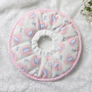 Puede incluir: Un collar de recuperaci&oacute;n acolchado para mascotas con un centro blanco y un exterior estampado en rosa y blanco. El patr&oacute;n incluye corazones rosas, animales de dibujos animados azules y lazos. El collar est&aacute; dise&ntilde;ado para evitar que las mascotas se laman o muerdan las heridas.