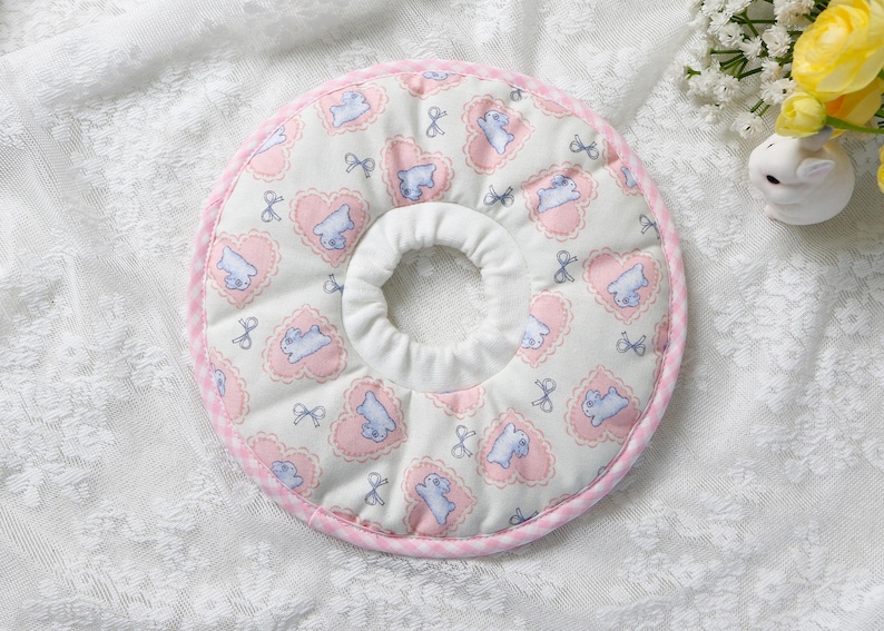 Puede incluir: Un collar de recuperaci&oacute;n acolchado para mascotas con un centro blanco y un exterior estampado en rosa y blanco. El patr&oacute;n incluye corazones rosas, animales de dibujos animados azules y lazos. El collar est&aacute; dise&ntilde;ado para evitar que las mascotas se laman o muerdan las heridas.
