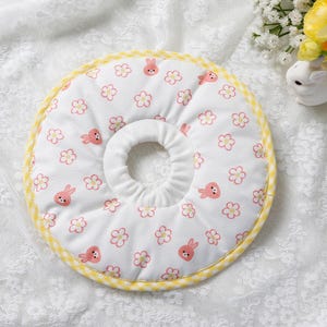 Puede incluir: Collar de recuperaci&oacute;n para mascotas blanco con un estampado de conejitos y flores rosas. El collar tiene un ribete de cuadros amarillos y un centro acolchado. El fondo es de tela de encaje blanco.