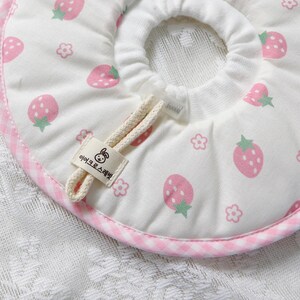 Puede incluir: Collar de recuperaci&oacute;n para mascotas blanco con un estampado de fresas rosas y un ribete de cuadros rosas. El collar tiene un cierre de cord&oacute;n y una peque&ntilde;a etiqueta con texto y un dibujo de conejo.