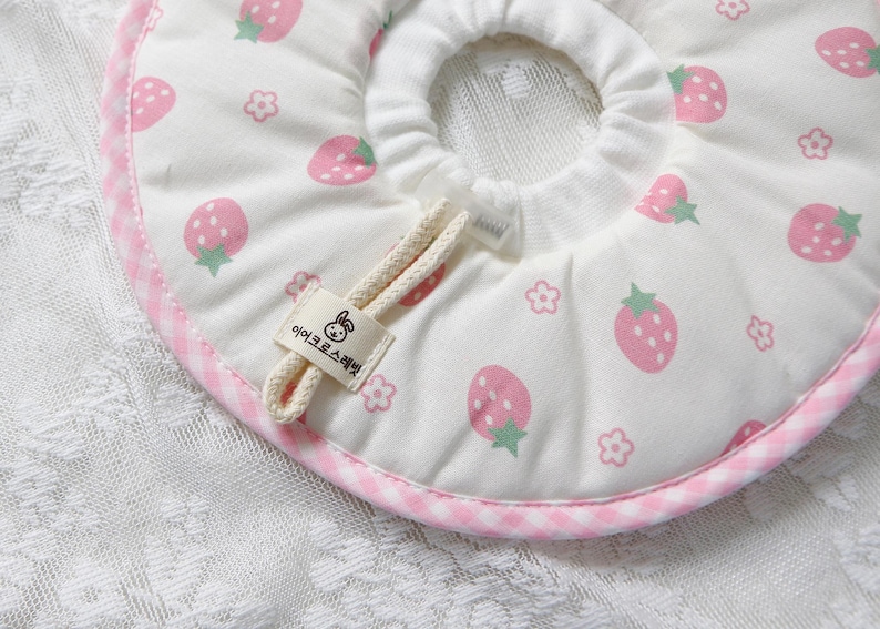 Puede incluir: Collar de recuperaci&oacute;n para mascotas blanco con un estampado de fresas rosas y un ribete de cuadros rosas. El collar tiene un cierre de cord&oacute;n y una peque&ntilde;a etiqueta con texto y un dibujo de conejo.